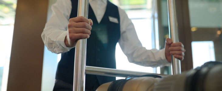 Cropped shot of young bellboy in uniform holding by metallic handles of baggage cart with suitcase of guests of modern hotel